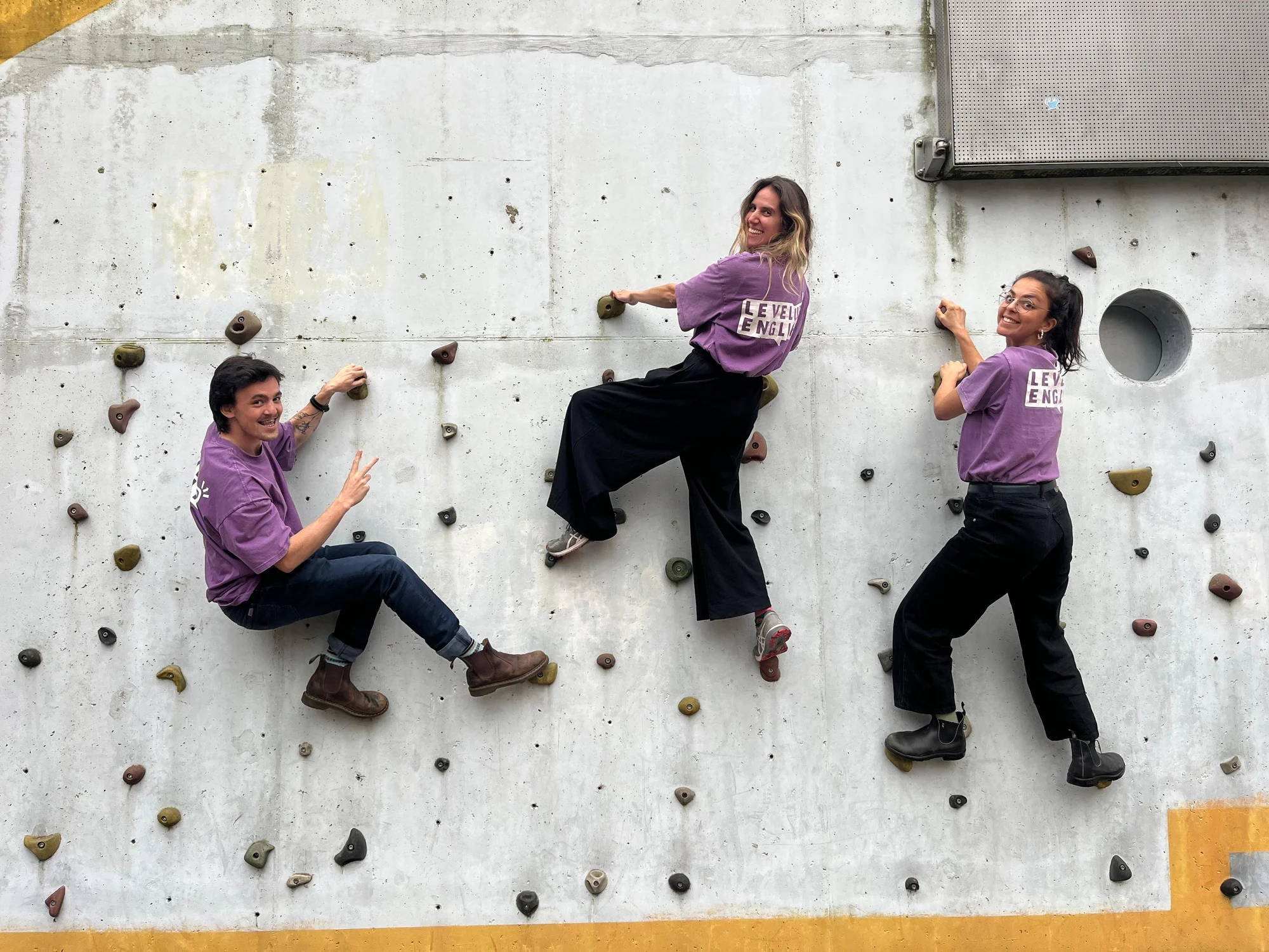 Three LevelUp trainers in purple LevelUp English shirts climbing a bouldering wall, laughing at the camera